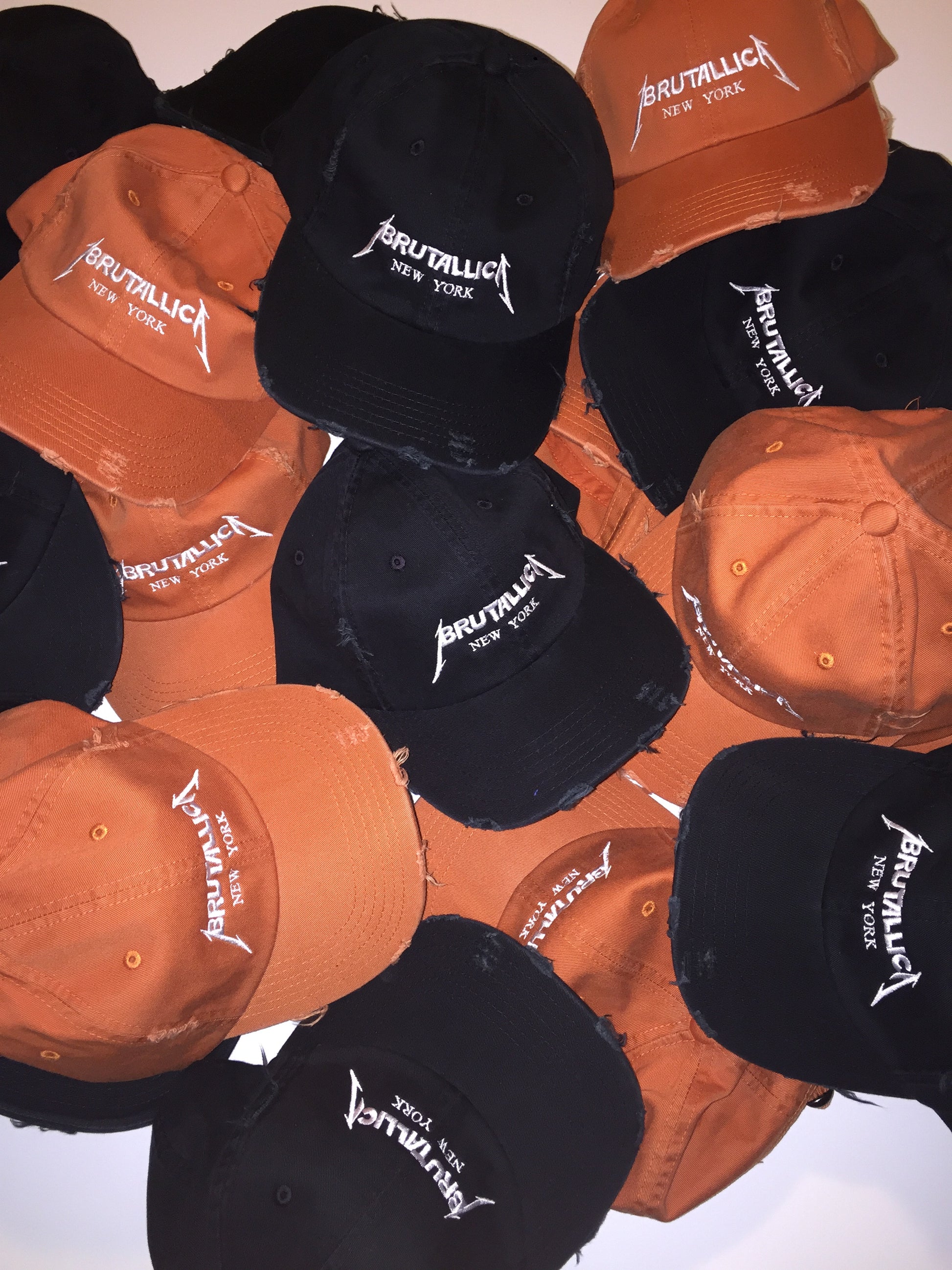 Brutallica distressed dad hats in black and burnt orange colorways with white embroidered logo pile of hats
