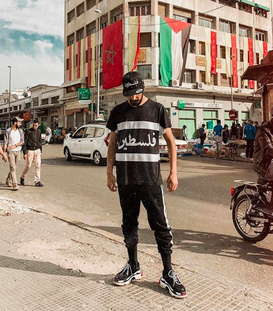 Palestine cut and sew shirt worn by male model in morocco in the streets with flags in the background, wearing brutallic shirt and hat with balenciaga shoes